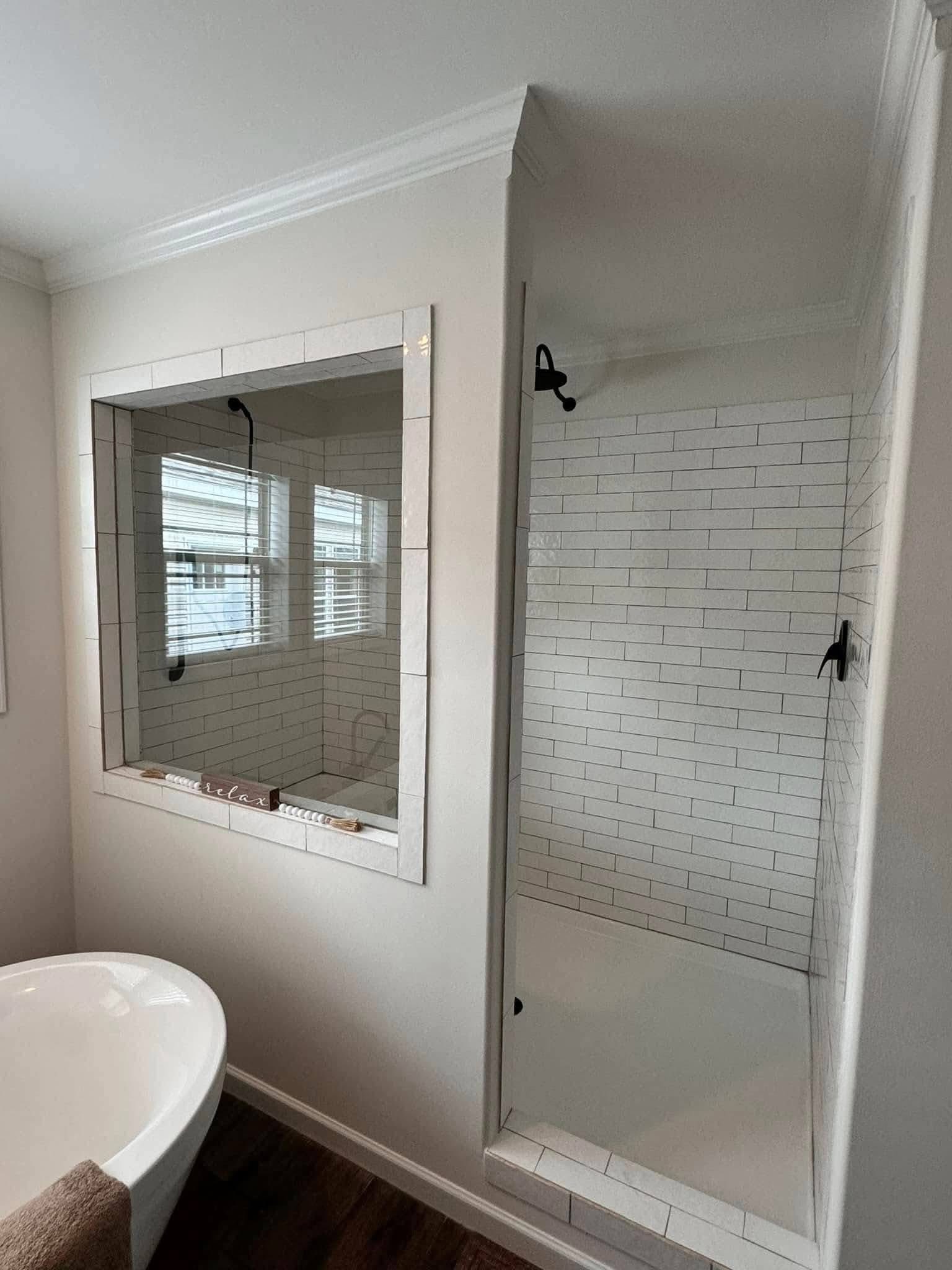 Modern bathroom with white subway-tiled walk-in shower, adjacent to a large mirror reflecting a window. A cozy bathtub and dark wood floor add warmth.