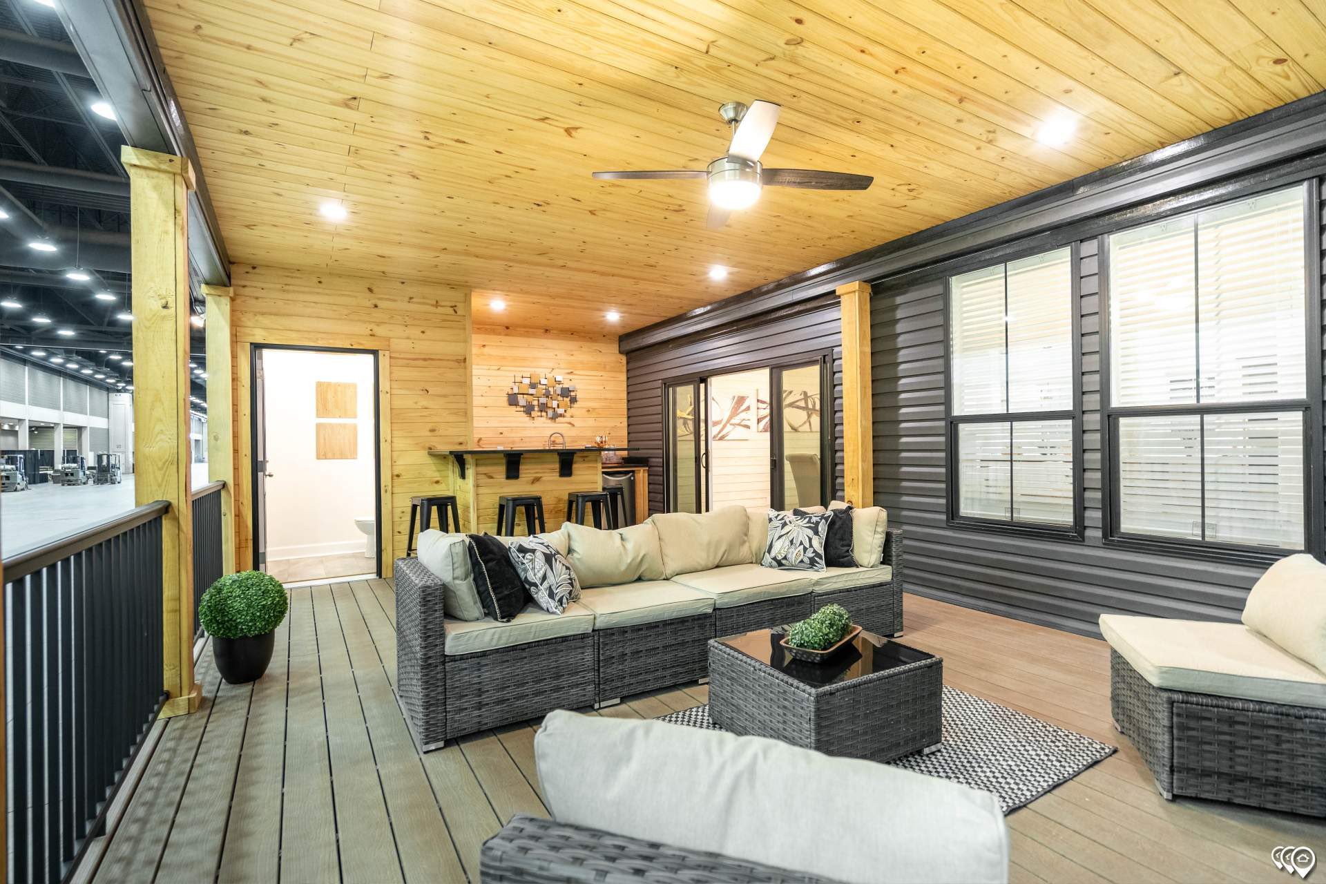A cozy indoor patio with wooden ceiling and floor, featuring a wicker sofa set with beige cushions, bar area with stools, and a ceiling fan above.