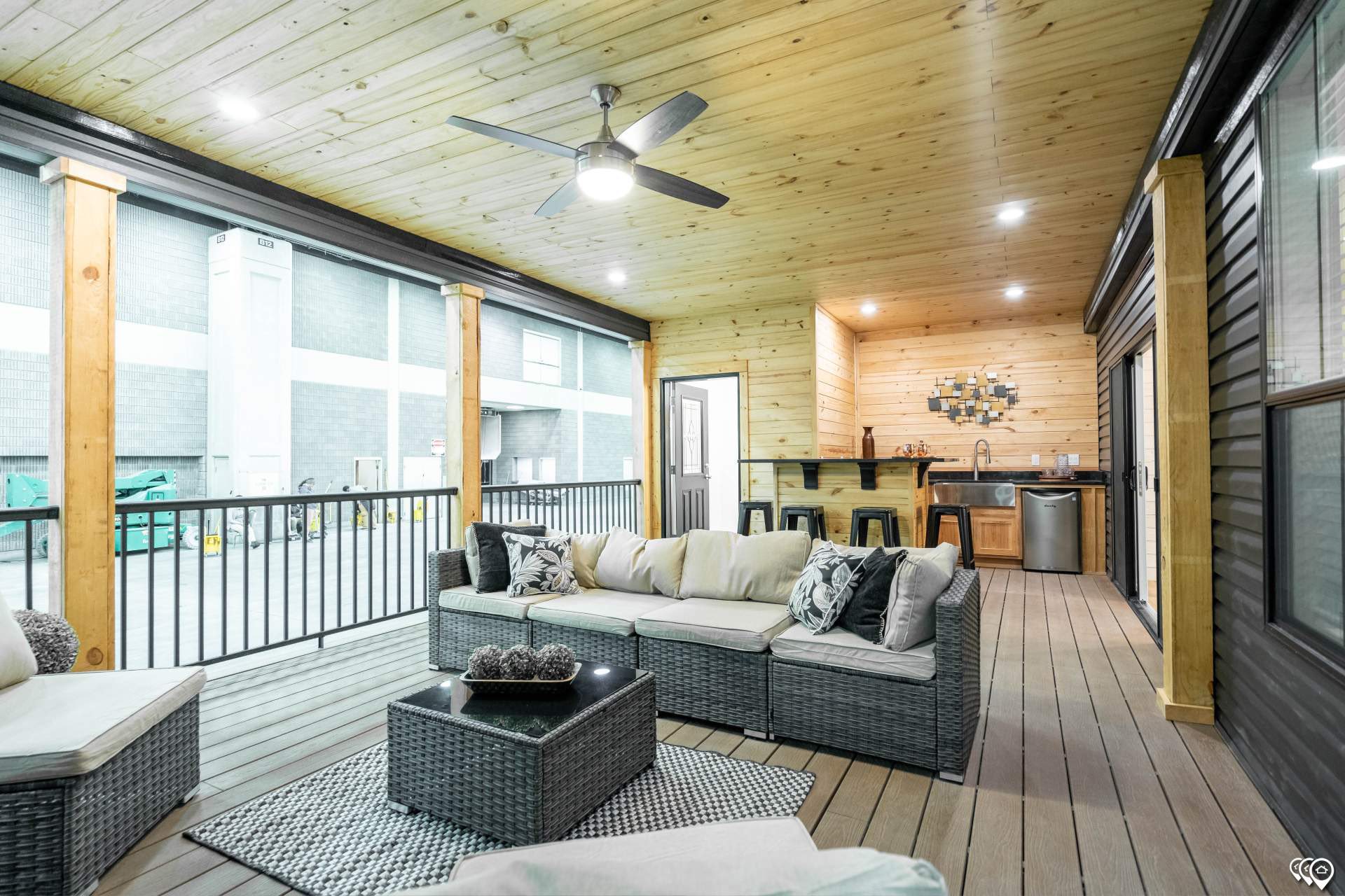 Covered patio featuring a modern wicker sofa set with cushions, a sleek ceiling fan, and a wooden bar area with stools, creating a cozy, inviting atmosphere.