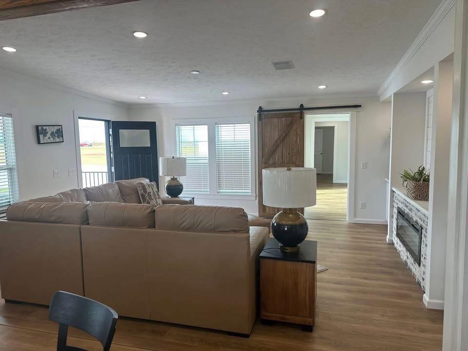 Cozy living room with tan sectional sofa, two table lamps, and wooden floors. Barn door, brick fireplace, and large windows create a warm ambiance.