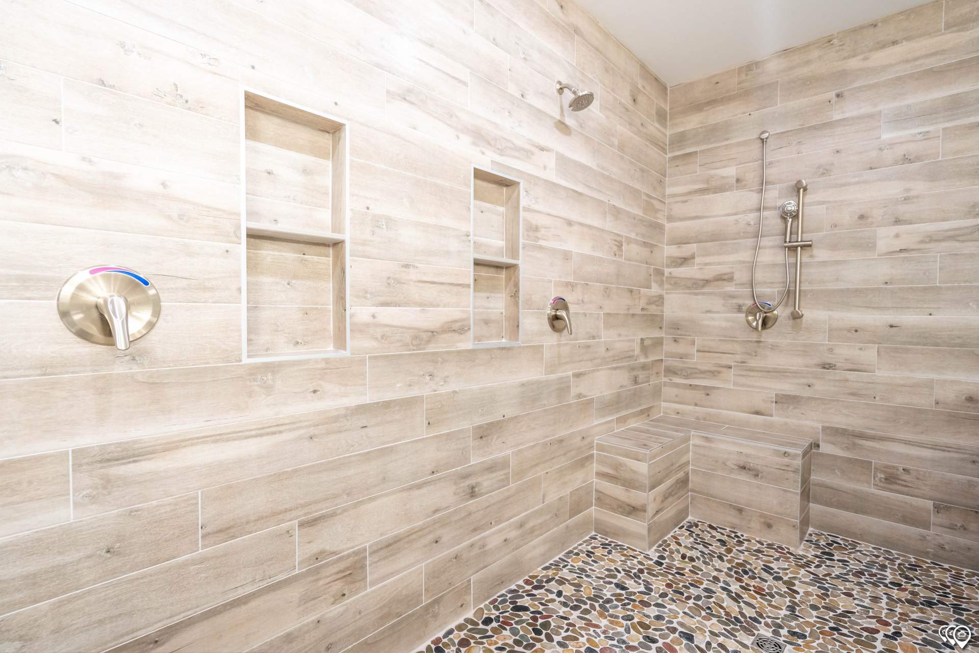 Modern shower with light wood-textured tile walls, pebble stone floor, chrome fixtures, built-in shelves, and a minimalist bench. Serene and spa-like.