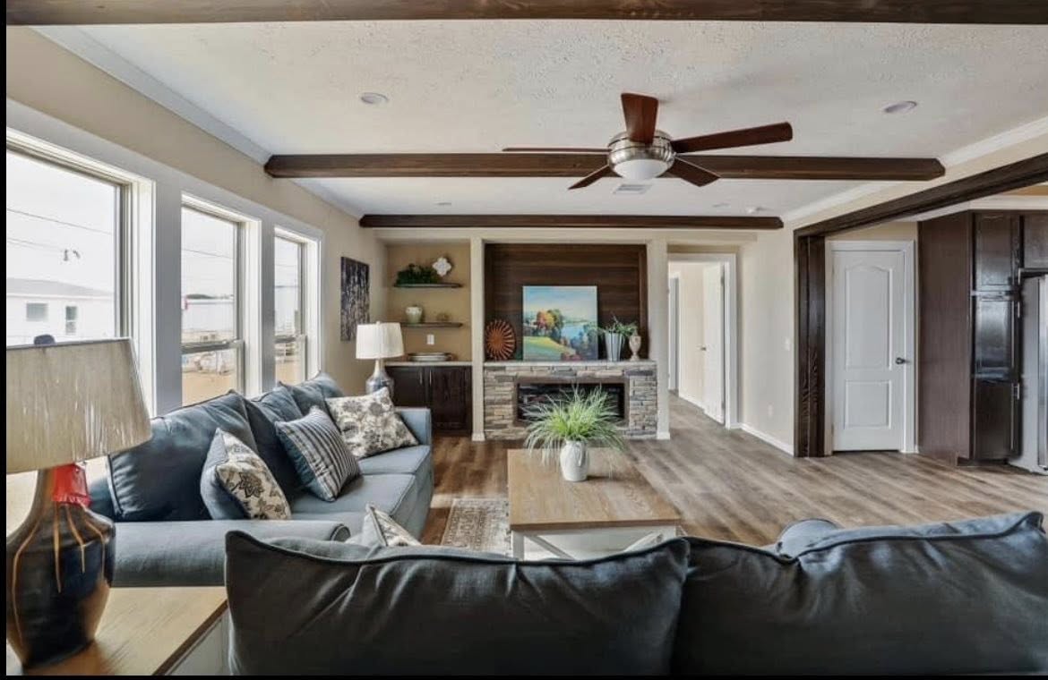 Spacious living room with wood flooring, cozy gray sofas, and decorative pillows. Large windows and a ceiling fan create an airy, inviting atmosphere.