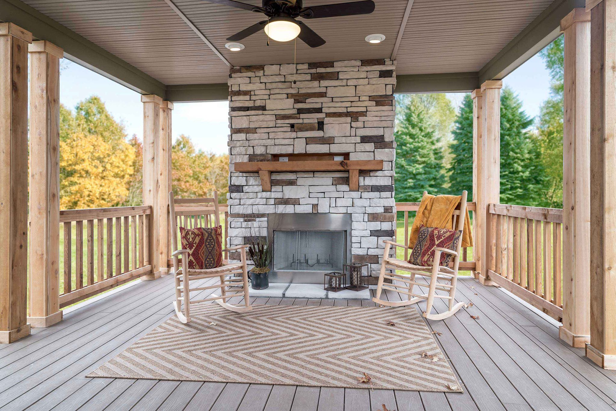 Covered porch with a stone fireplace and two wooden rocking chairs on a chevron-patterned rug. Surrounded by autumn trees, creating a cozy, inviting atmosphere.