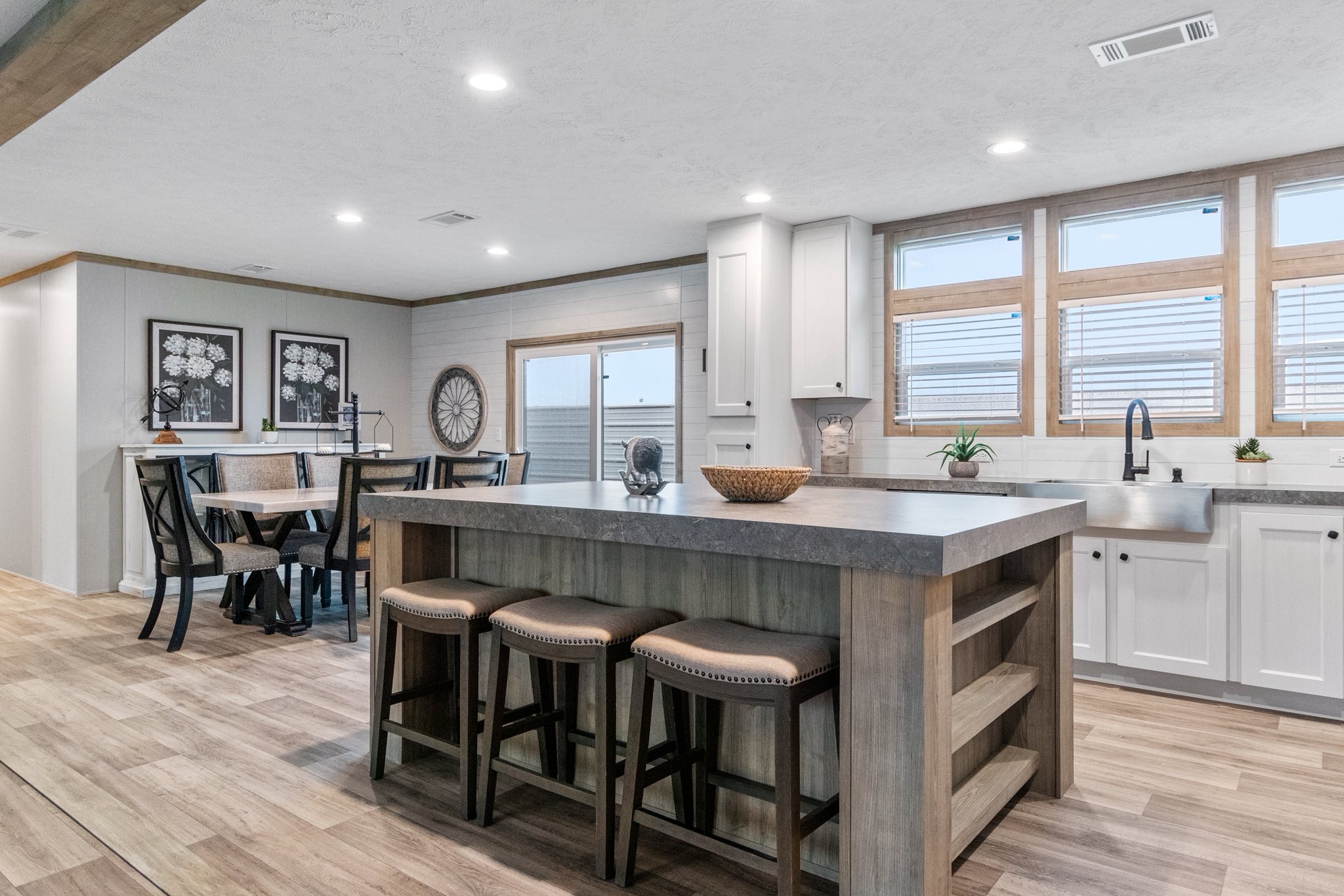 Modern kitchen and dining area with wood flooring. Features a large island with three stools, a sink with windows above, and a table with chairs. Bright and inviting.