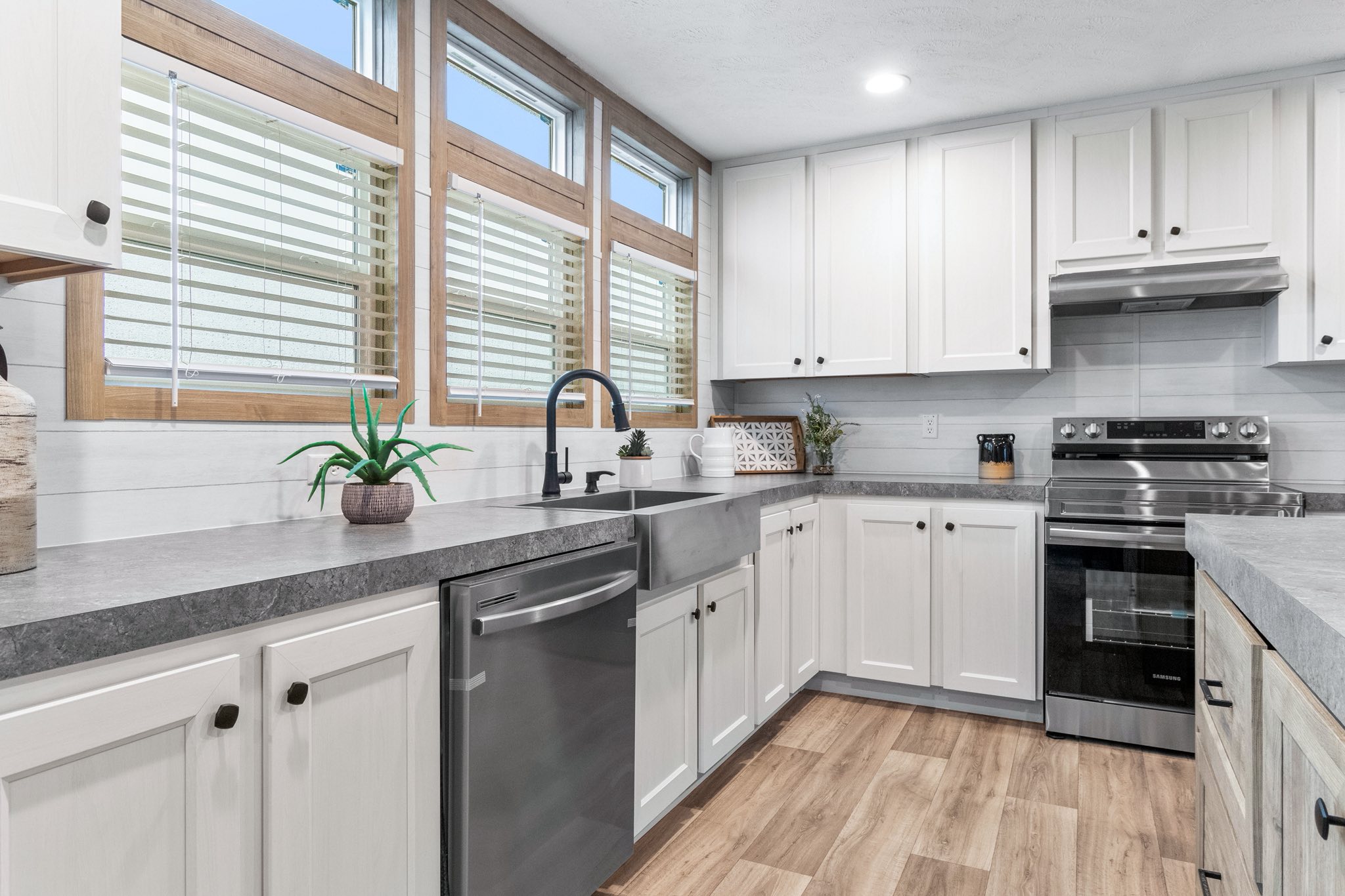 Bright kitchen with white cabinets, stainless steel appliances, and a farmhouse sink. Large windows with blinds let in natural light onto the wooden floor.
