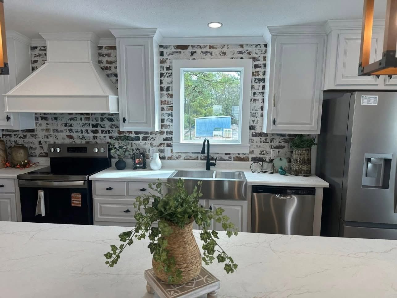 Modern kitchen with white cabinetry and brick backsplash. Central window with natural light. Stainless steel appliances. Green plant on marble counter.