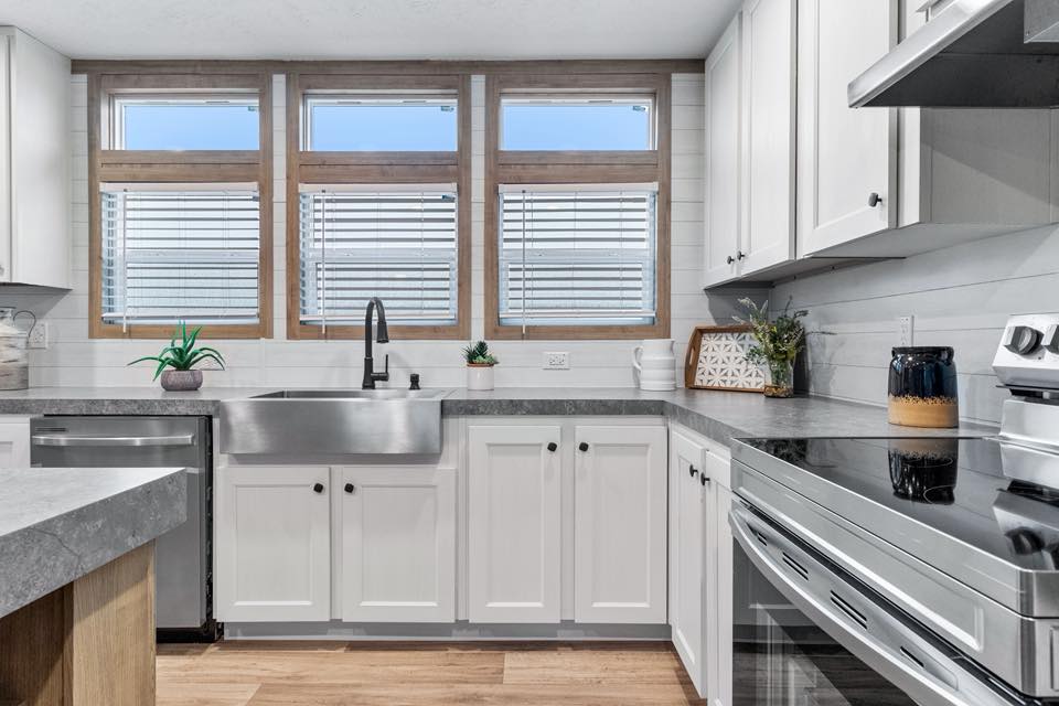 Modern kitchen with white cabinets, stainless steel appliances, and a farmhouse sink. Three large windows provide natural light, creating a bright and airy atmosphere.