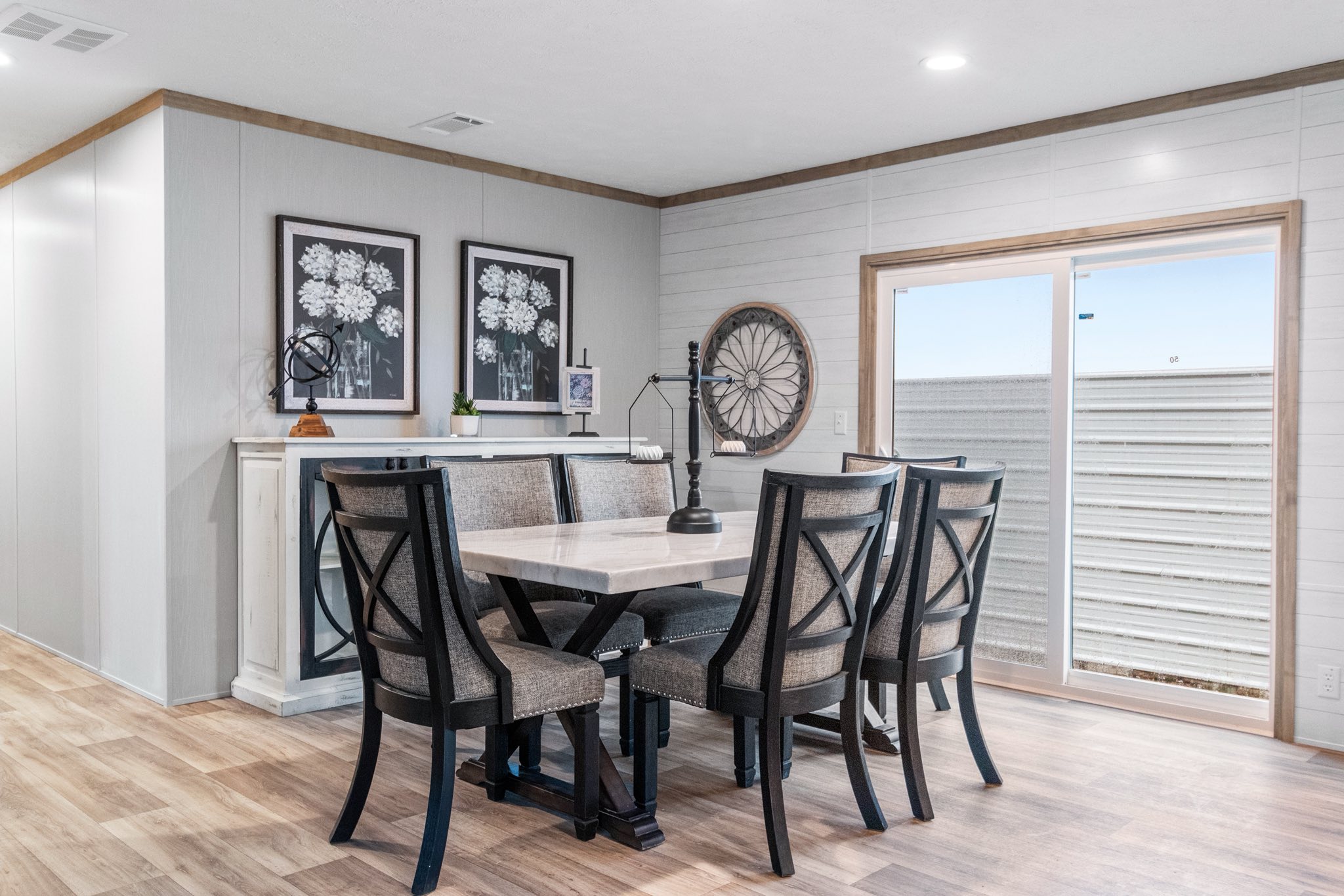 A modern dining room with a light wood floor features a rectangular table and six cushioned chairs with dark trim. Two floral artworks hang on a sideboard, accompanied by a lamp, candlestick, and decor. A large circular piece is mounted on a white panel wall beside a sliding glass door, letting in natural light. The space feels cozy and contemporary.