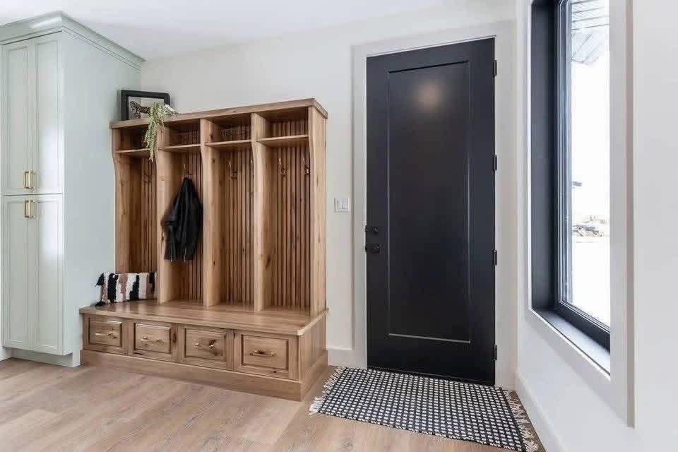 Wooden mudroom entryway with cubbies, coat hooks, and storage drawers. Black door and window, light wooden floor, and a black-and-white rug. Cozy and organized.