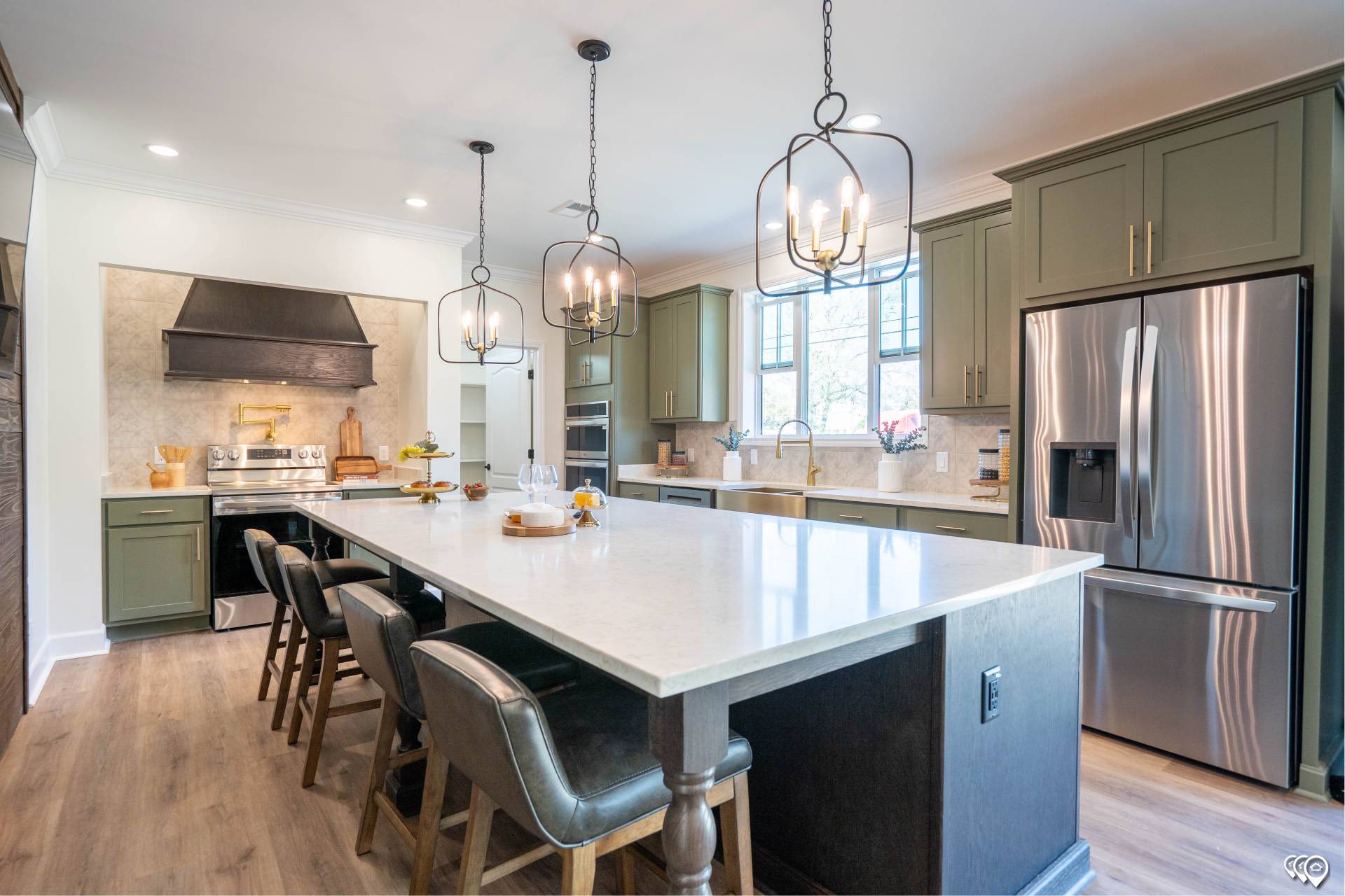 Spacious modern kitchen with a large white island, dark leather chairs, green cabinetry, stainless steel appliances, and elegant pendant lighting.