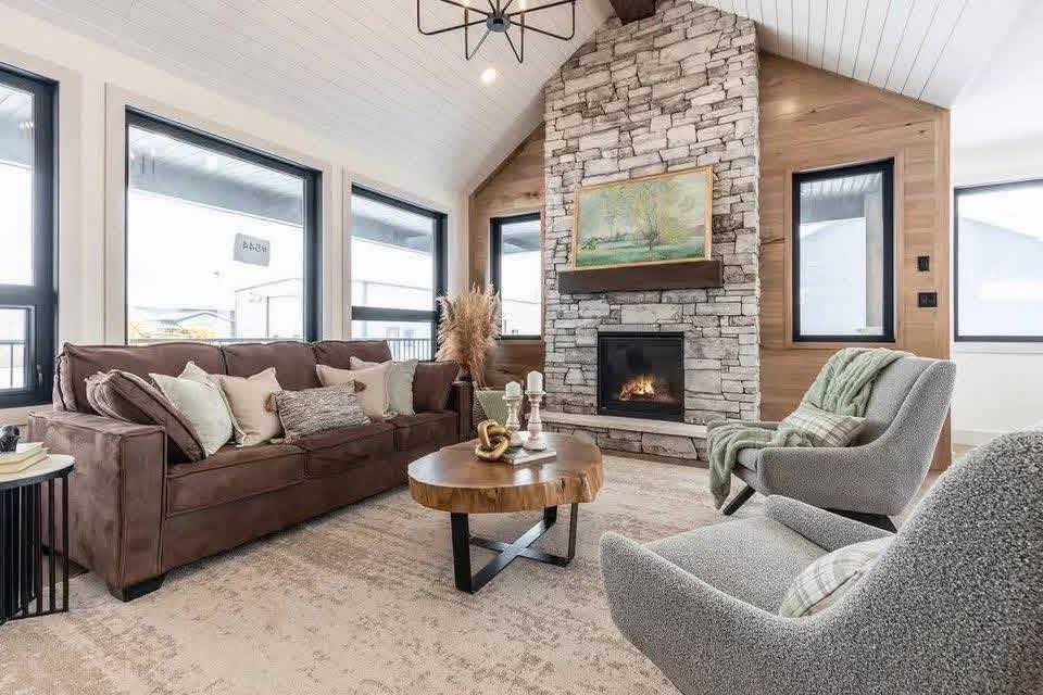 Cozy living room with stone fireplace, brown sofa, and gray armchairs. Wooden accents and large windows create a warm, inviting atmosphere.