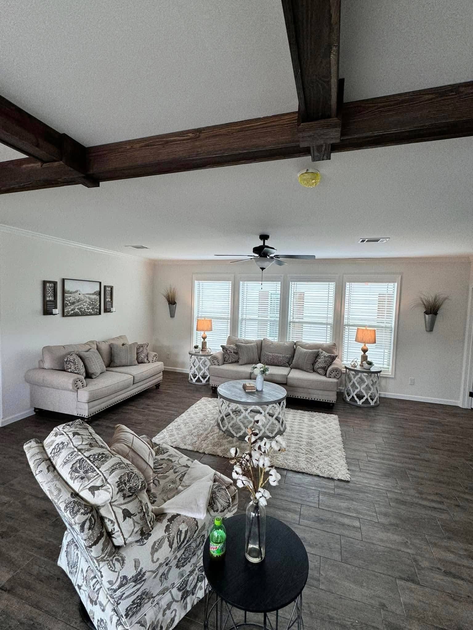 Spacious living room with light beige sofas, floral armchairs, and a round table. Large windows, wooden beams, and decor create a cozy, elegant ambiance.