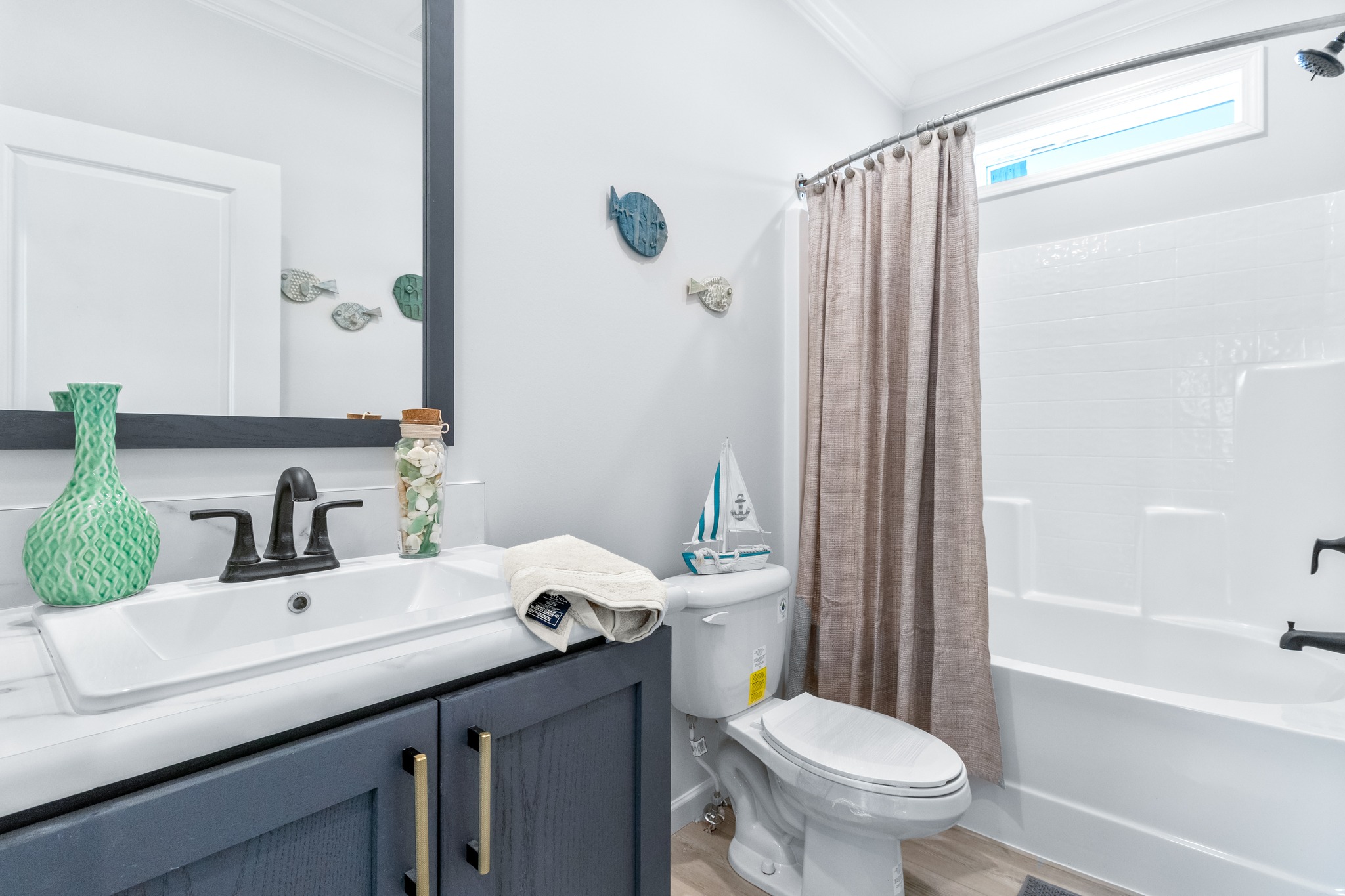 Bright bathroom with nautical decor, featuring a dark sink vanity, seashell accents, a model sailboat on the toilet, and a beige shower curtain.