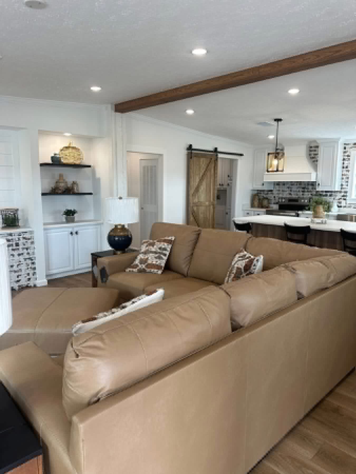 A cozy living room features a large tan sectional sofa with patterned cushions. A lamp sits on a side table. The open kitchen has a farmhouse style with white cabinets, a wooden sliding barn door, and industrial light fixtures, creating a warm, inviting atmosphere.