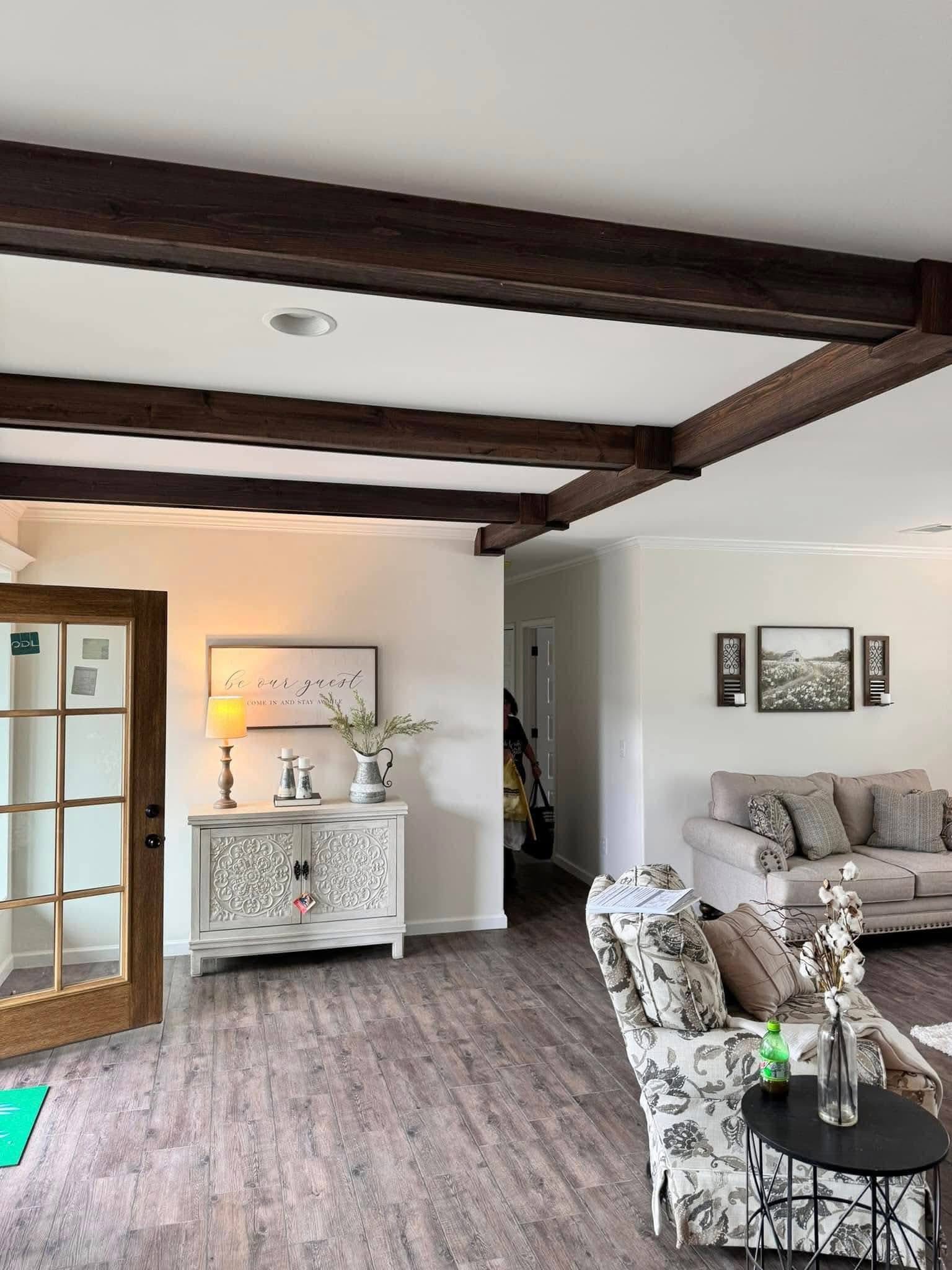 Living room with exposed dark wooden beams and wooden floor. A cozy sofa features patterned pillows, a floral chair, and a side table with decor.