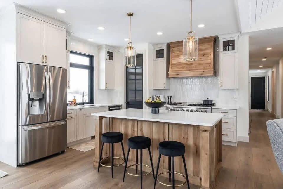 Modern kitchen with a large stainless steel fridge, white cabinets, and a central island with wooden accents and three black stools. Cozy and elegant ambiance.