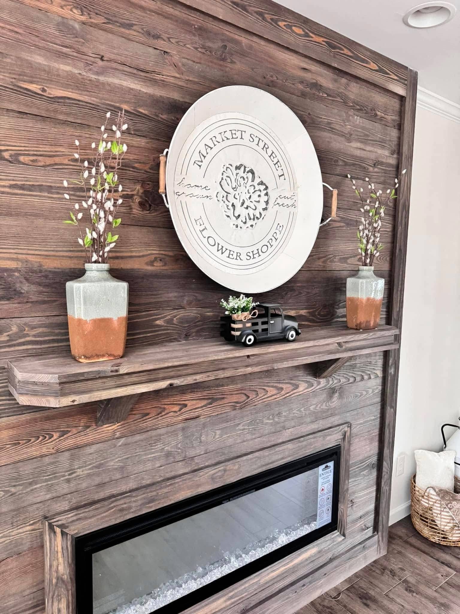 Rustic wooden wall with a circular Market Street Flower Shoppe sign. Two vases with white flowers flank a toy truck on a shelf above a modern fireplace.