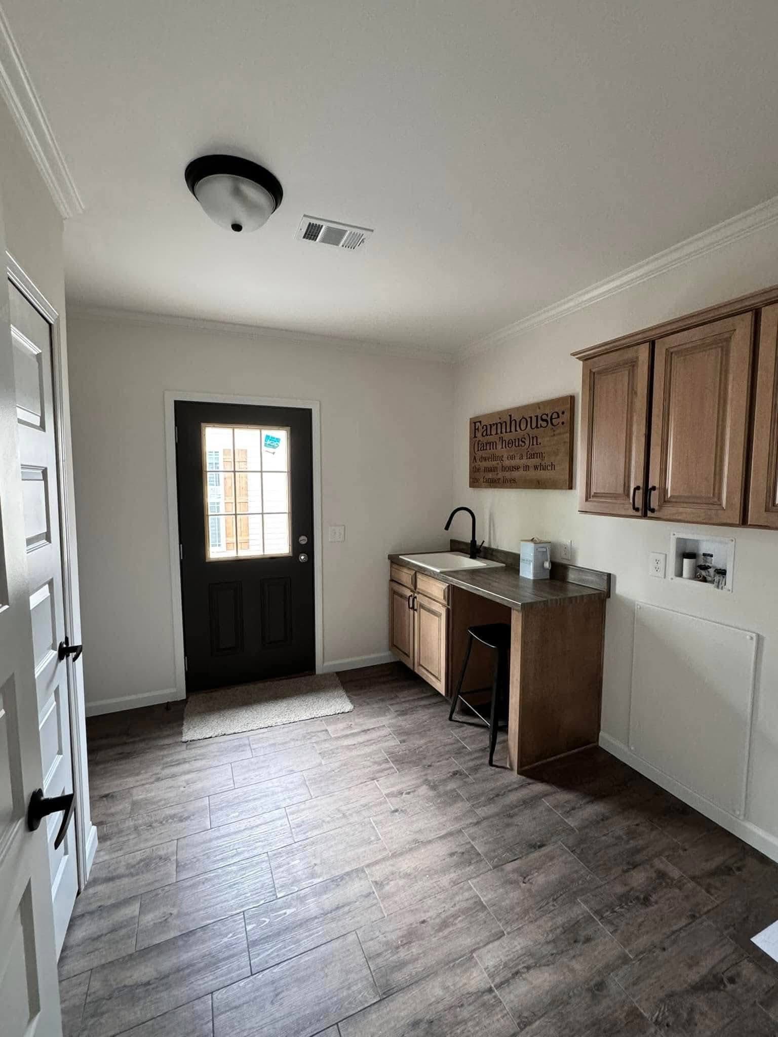 A small, rustic-style laundry room with wood flooring, a black door, and wooden cabinets. A sink area is adorned with a farmhouse-themed sign.
