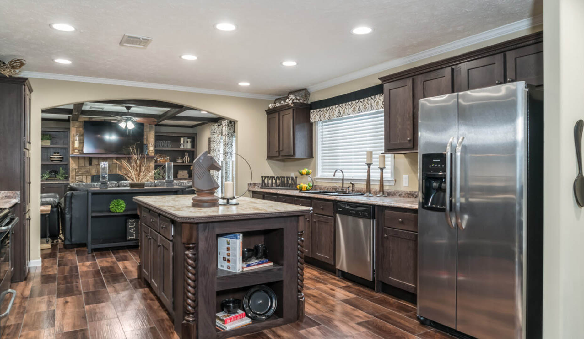 Modern kitchen with dark wooden cabinets, stainless steel appliances, and an island. Warm lighting on wooden floors creates a cozy atmosphere.