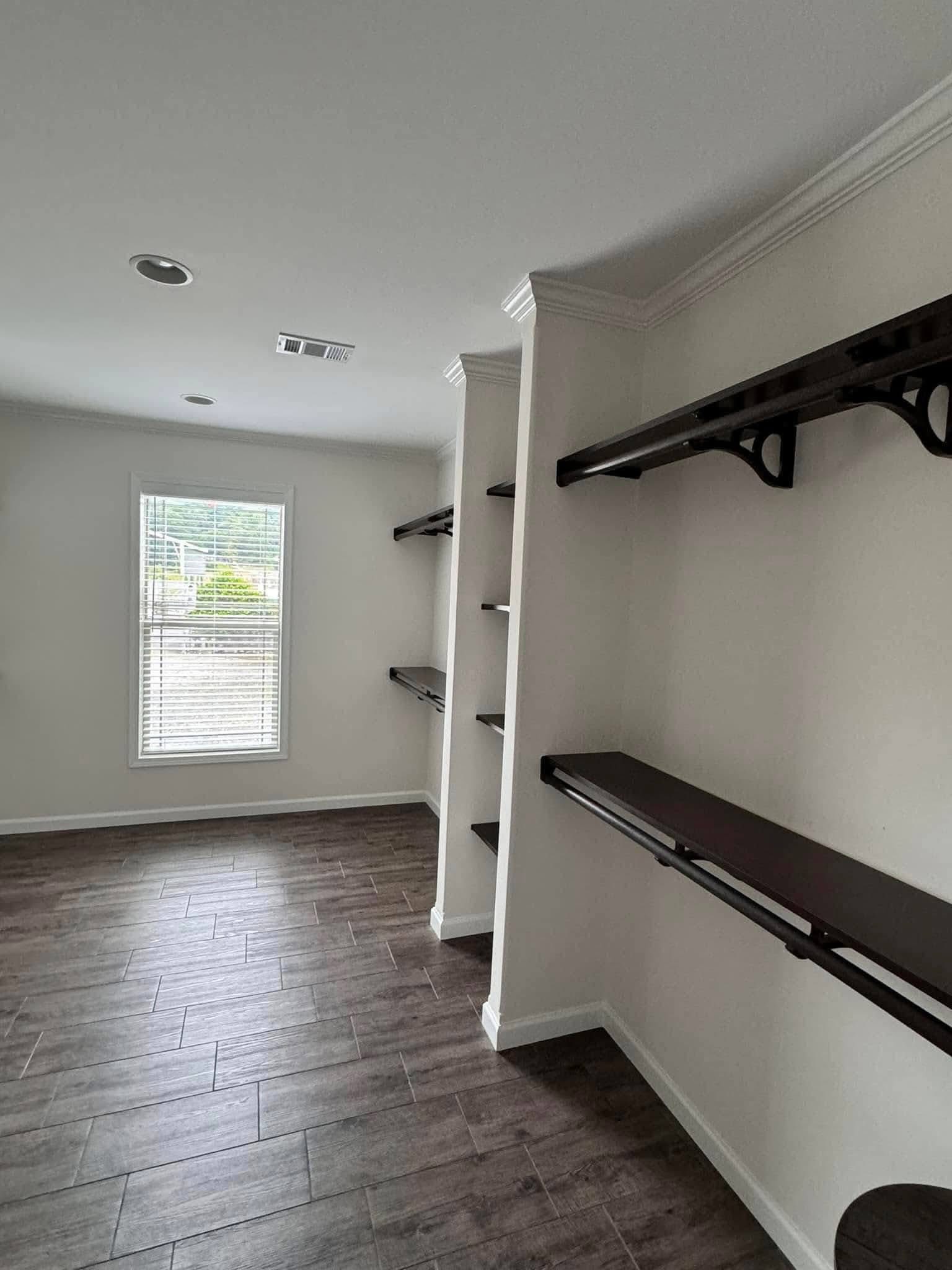 Bright walk-in closet with wooden shelves and rods on cream walls. Large window illuminates dark tile flooring, creating a spacious, organized feel.