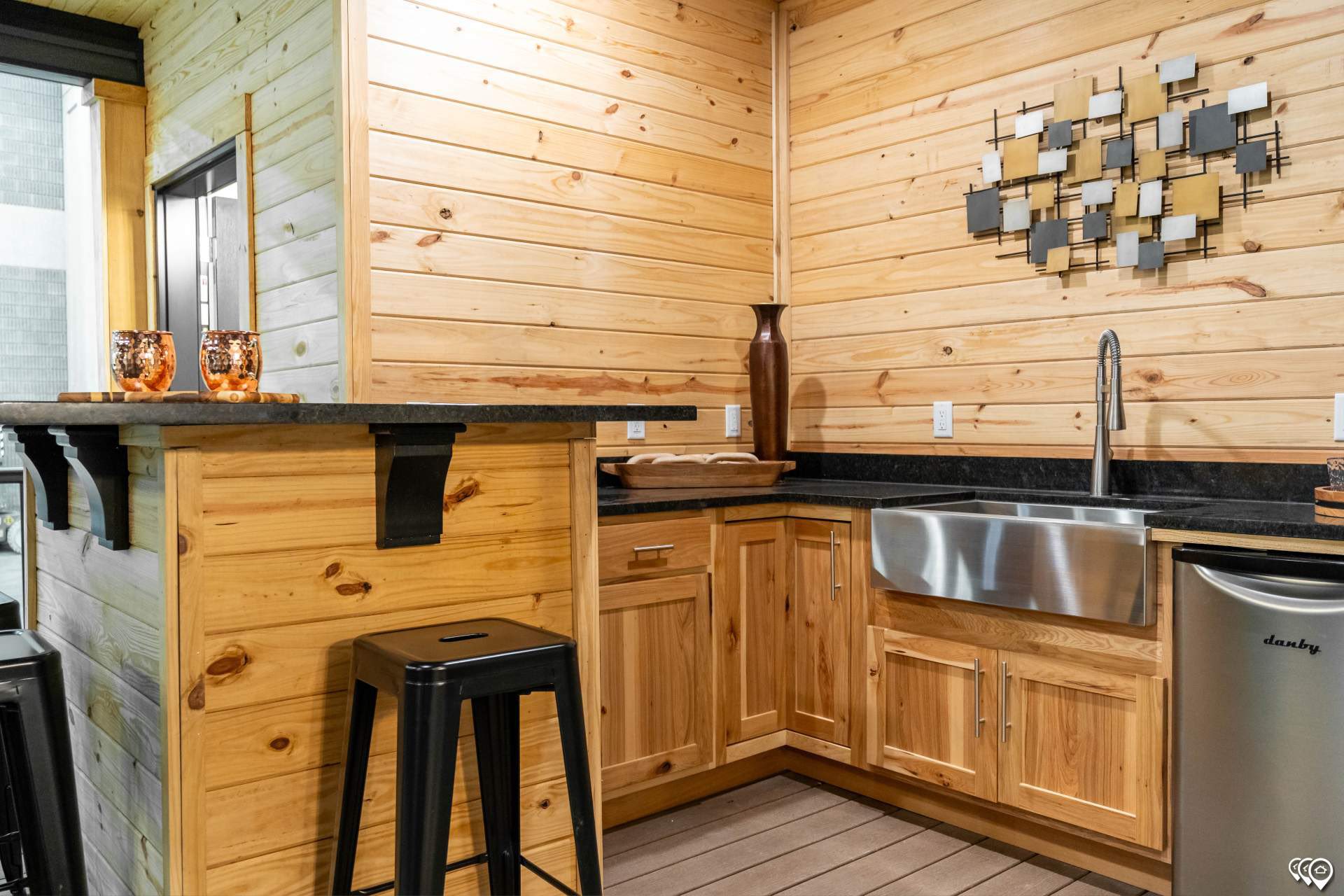 Cozy kitchenette with light wood paneling, black countertop, and stainless steel sink. Features barstools, abstract wall art, and warm, inviting tones.