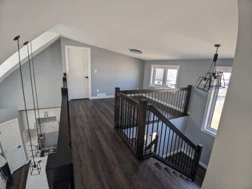 A modern, bright hallway with dark wood flooring, stairs with black railing, and large windows. Minimalist light fixtures hang from a high ceiling.