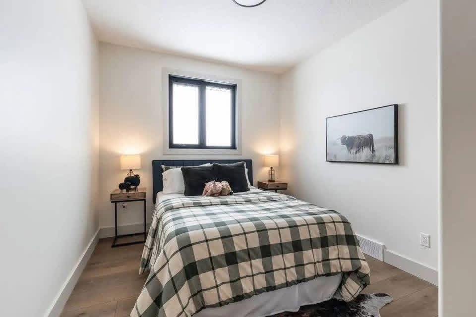 Cozy bedroom with a plaid-covered bed centered between two nightstands with lamps. A framed cow photo hangs on the right wall. Warm and inviting.