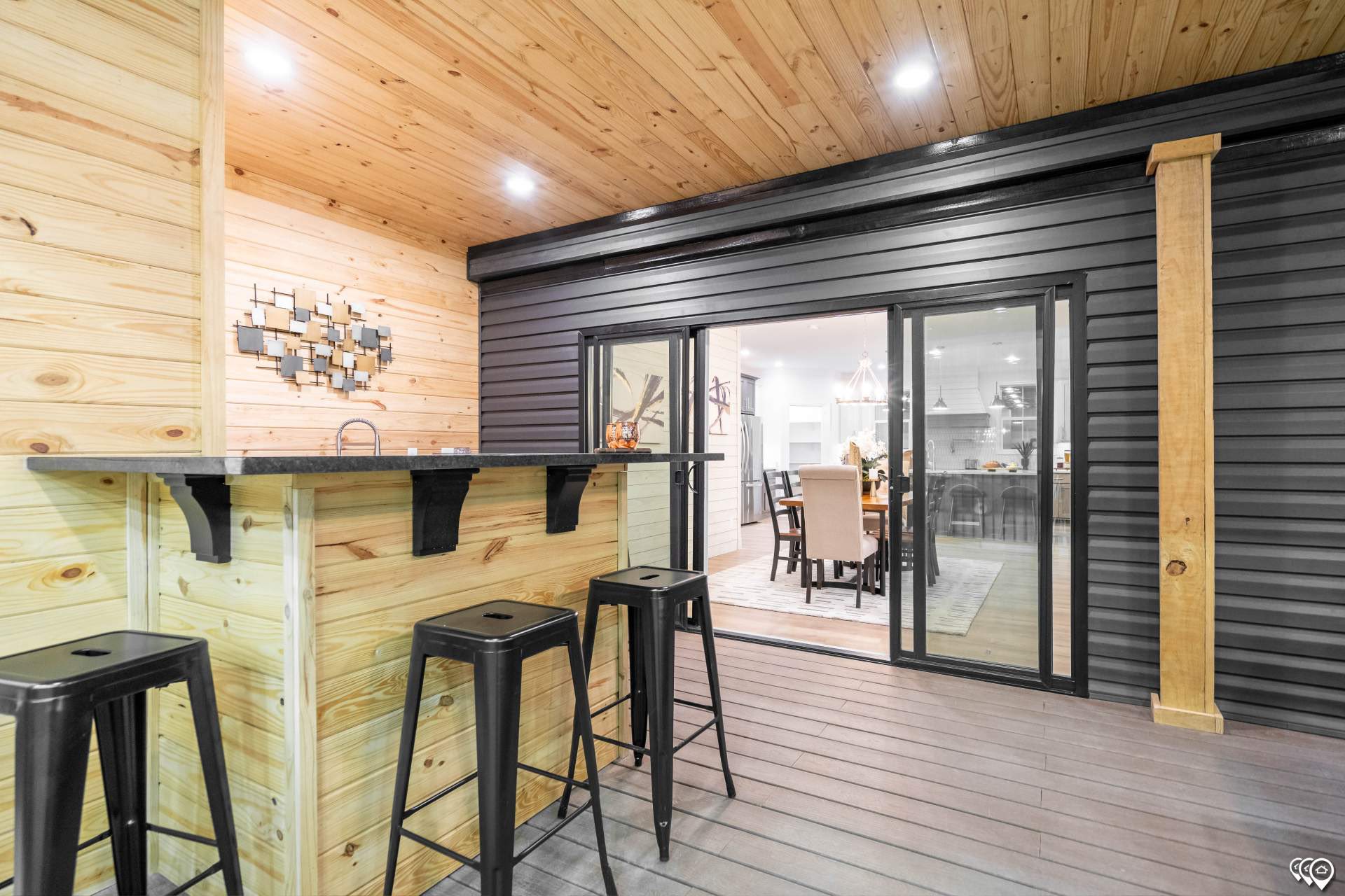 Stylish outdoor bar with wood paneling, three black stools, and a decorative wall piece. Glass doors open to a warmly lit dining room, creating a cozy ambiance.
