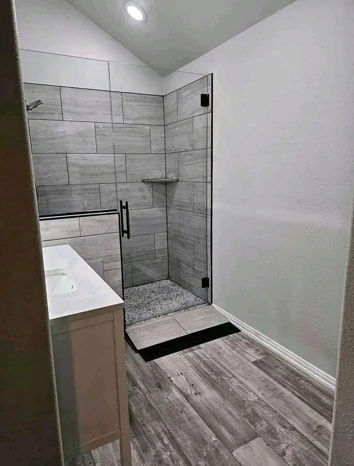 Modern bathroom with a gray tiled walk-in shower, glass door, and sleek black handle. White vanity and wood-like flooring create a clean, minimalistic look.