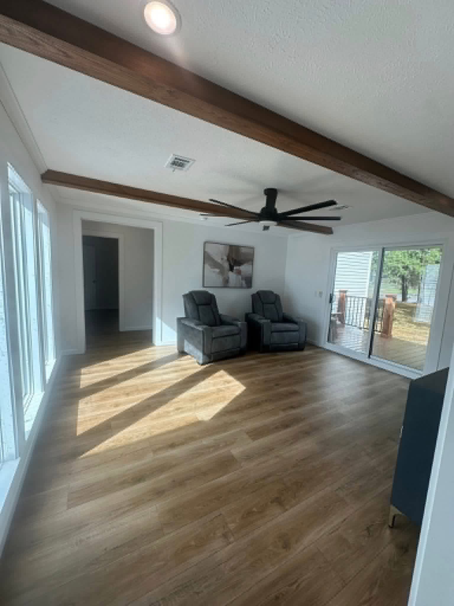 Bright room with wood flooring, two gray recliners, ceiling fan, and art piece. Large windows and sliding door open to a deck, creating an airy feel.