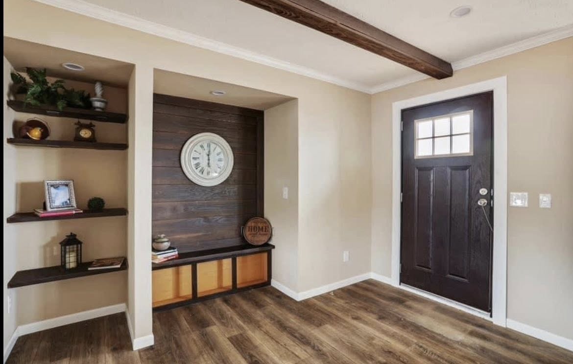 Warm entryway with beige walls, wood flooring, and a dark wooden door. A wall clock is set against dark paneling, with shelves holding decor items. Cozy ambiance.