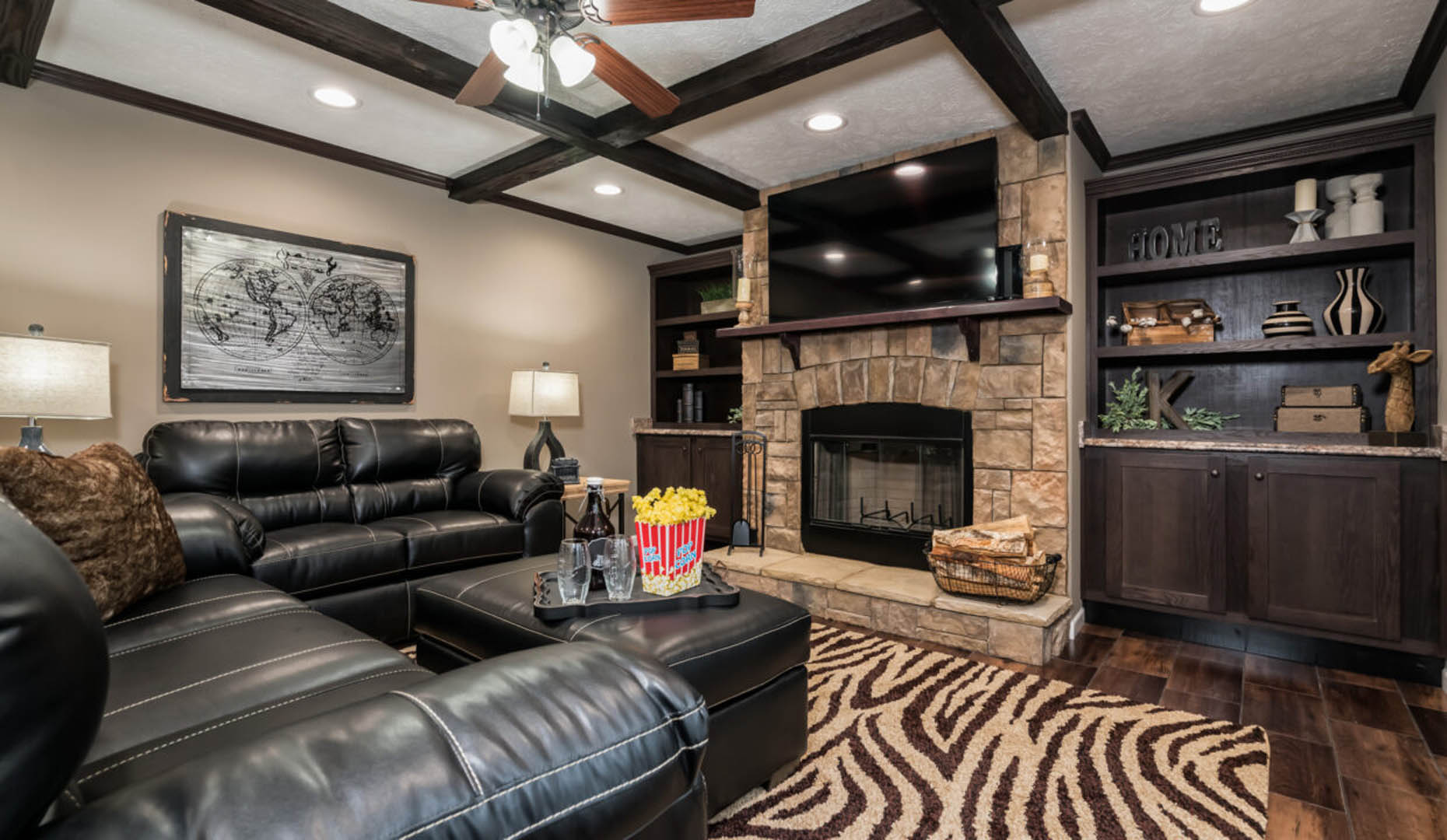 A cozy living room with dark leather sofas, a stone fireplace, and a TV above it. A zebra-patterned rug and shelves with decor add warmth and style.