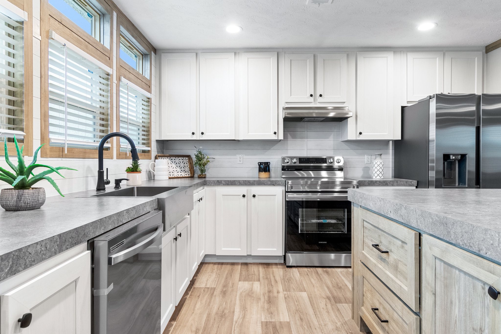 Modern kitchen with white cabinets, stainless steel appliances, and gray countertops. Natural light streams through large windows, highlighting a sleek, airy design.