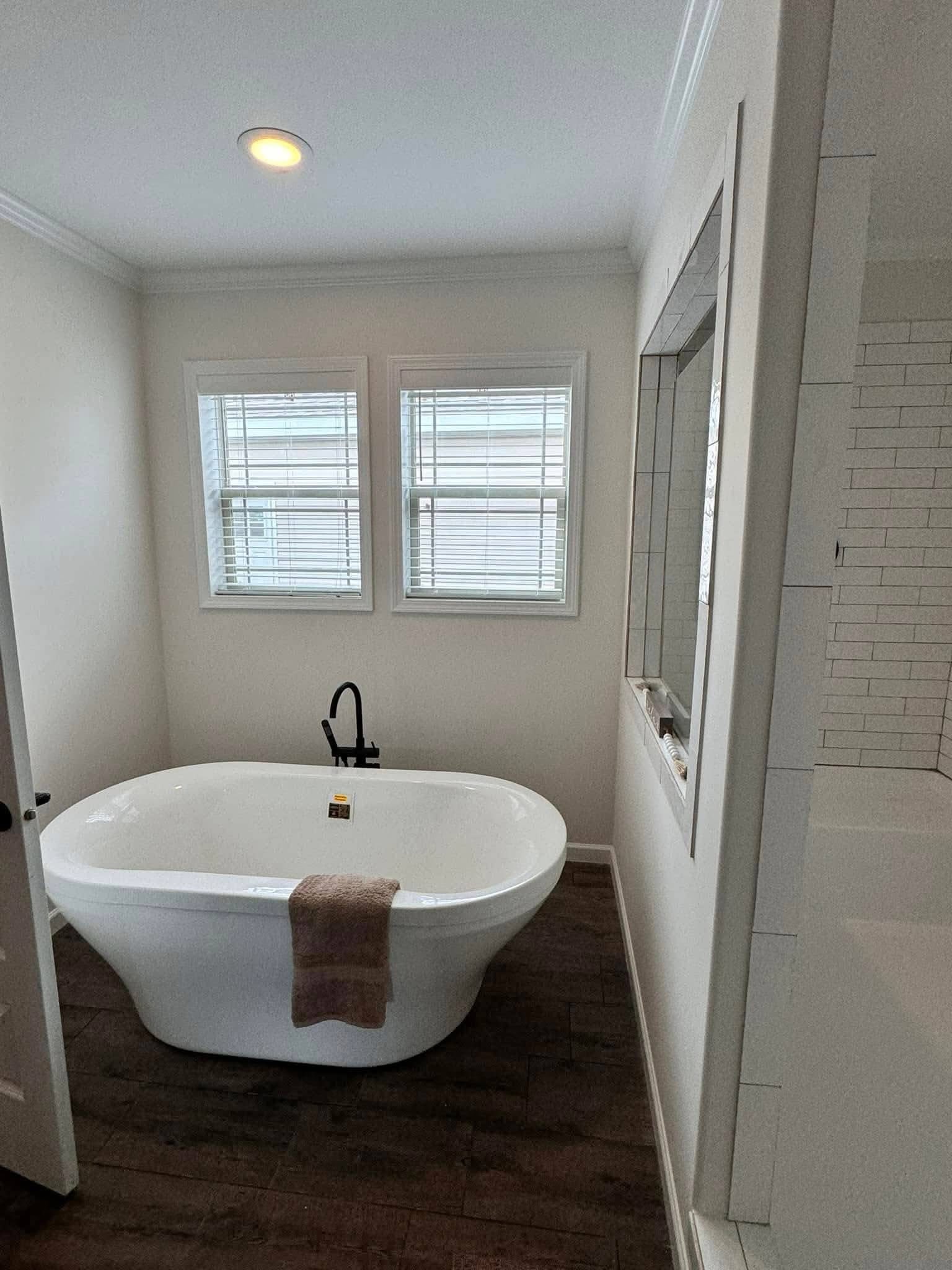 Freestanding white bathtub with a brown towel on it, set on dark wood flooring. Two windows with blinds above, creating a serene, minimalist atmosphere.
