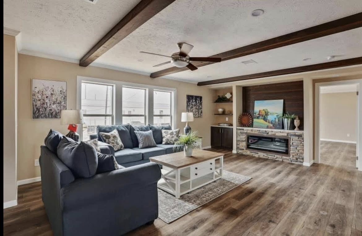 Spacious living room with a blue sectional sofa, wooden coffee table, ceiling fan, and stone fireplace. Warm tones and natural light create a cozy atmosphere.