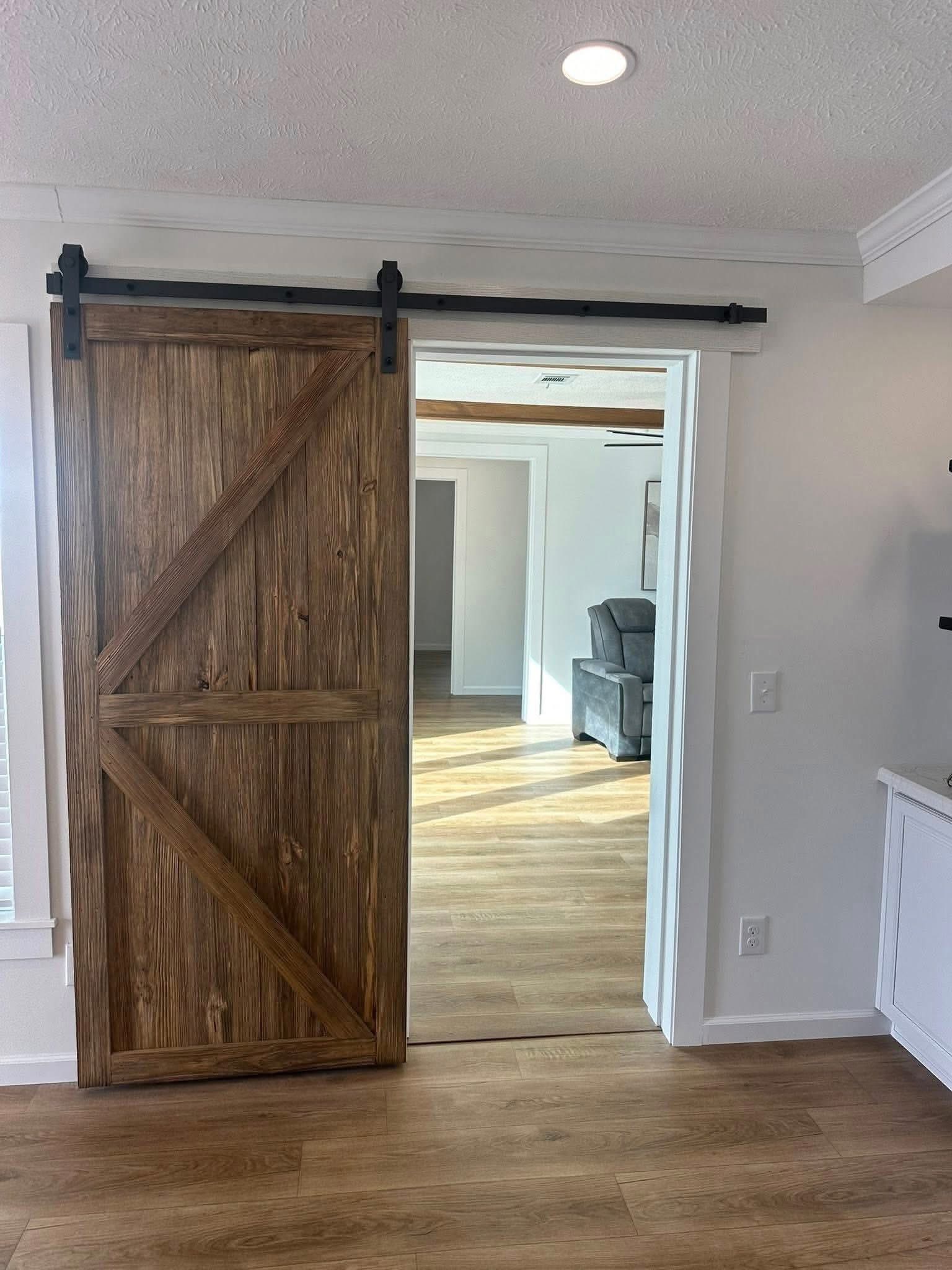 A rustic wooden sliding barn door partially open, revealing a bright room with wood flooring and a gray armchair. The setting feels cozy and inviting.