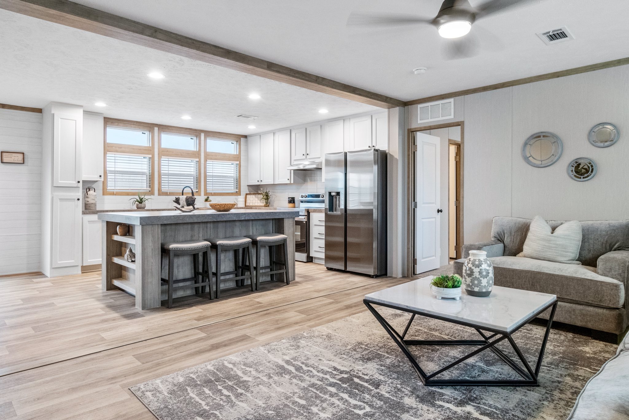 Modern open-concept living room and kitchen, featuring light wood floors, a gray sofa, sleek kitchen island with barstools, and stainless steel appliances.