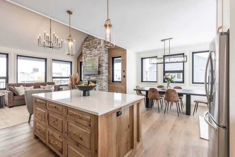 Modern open-plan kitchen and living room with a wooden island, pendant lights, stone accent wall, cozy seating, and a dining area with brown chairs.