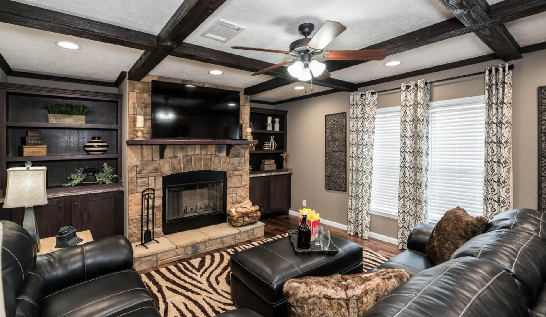 Cozy living room with dark leather sofas and a zebra-striped rug. Features a stone fireplace, built-in wooden shelves, and patterned curtains under a ceiling fan.