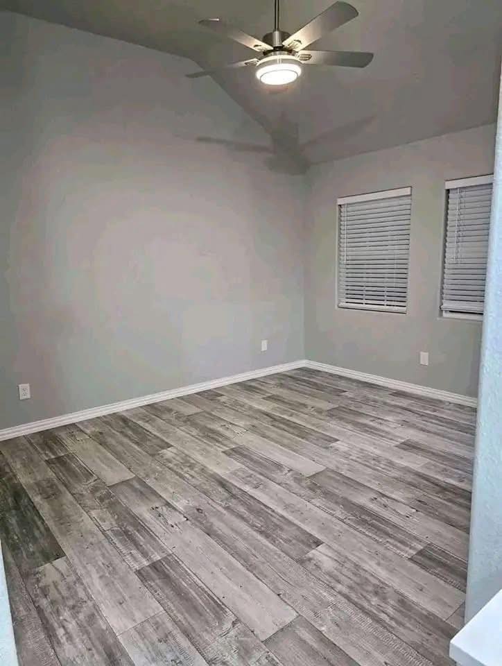 Empty room with gray walls and wood-look tile floor. Two windows with closed blinds illuminate the space, and a ceiling fan with a light fixture is on.