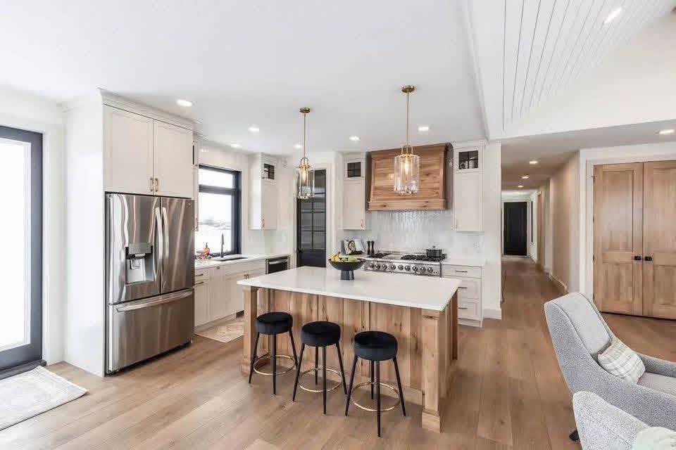 Modern kitchen with white cabinetry, stainless steel appliances, and a central island with three black stools. Light wood floors and pendant lights add warmth.