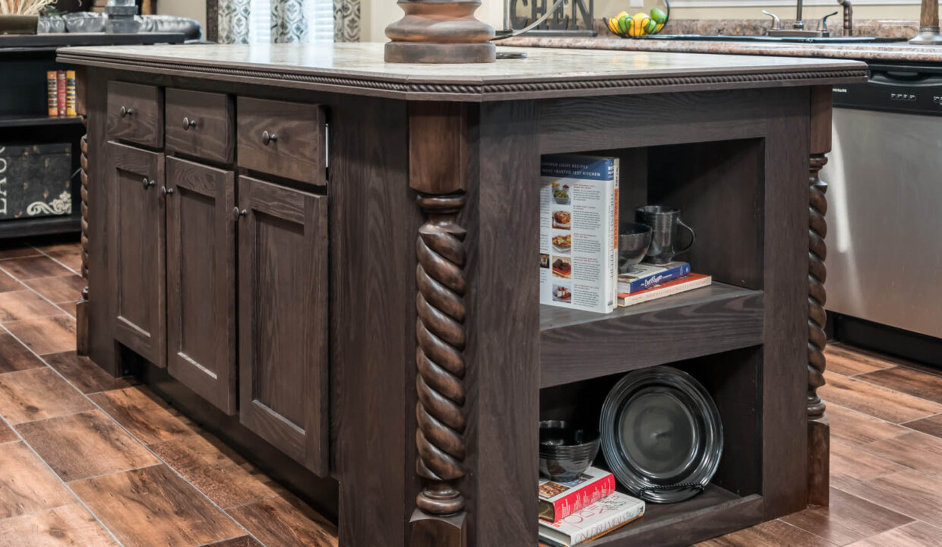 A rustic dark wood kitchen island with twisted column details, storage shelves holding cookbooks and dishes, and a warm, inviting atmosphere.