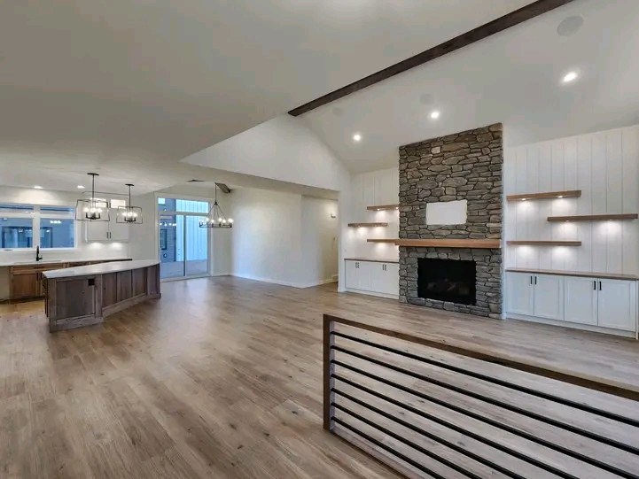 Spacious modern living room with wood floors, a stone fireplace, built-in shelves, and a kitchen island. Bright lighting creates a cozy ambiance.
