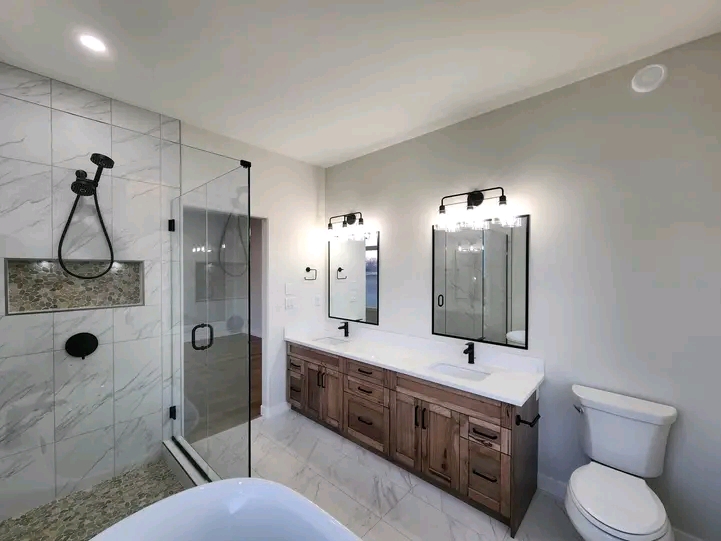 Modern bathroom with a glass shower, dual sink vanity with wooden cabinets, two mirrors, elegant lighting, and a white toilet, conveying a clean, sleek look.