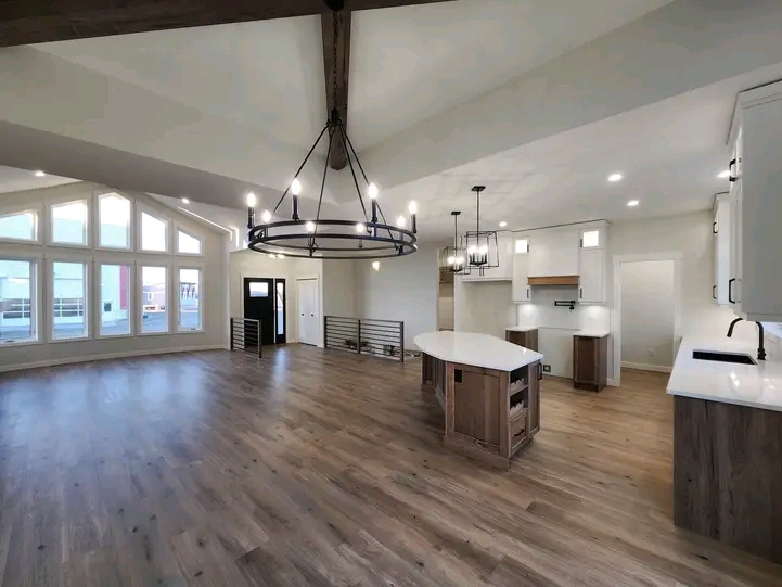 Spacious, modern open-plan kitchen and living area with vaulted ceilings, large windows, wooden floors, and a central chandelier. Bright and inviting ambiance.
