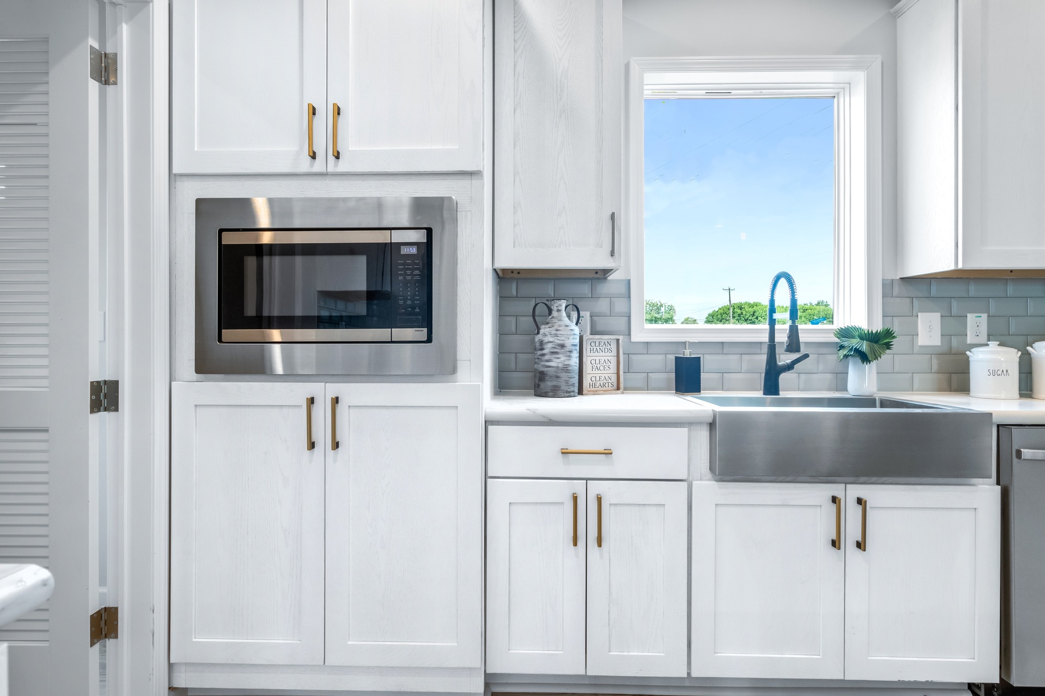 Bright kitchen with white cabinets and gold handles, featuring a built-in microwave, stainless steel sink, and window with a clear blue sky view.