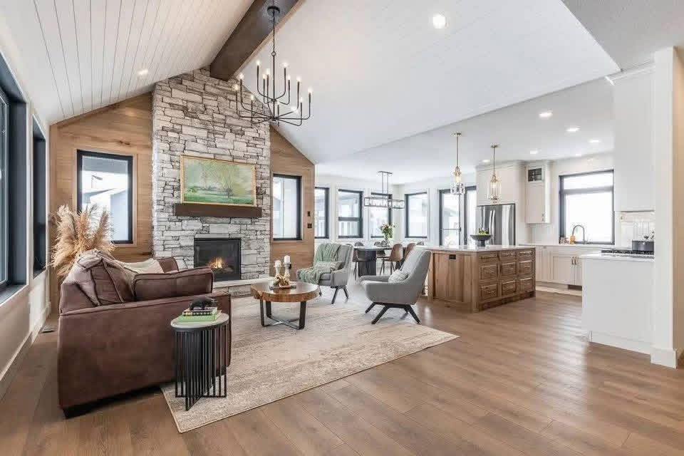 Modern open-concept living room with vaulted ceiling and rustic stone fireplace. Cozy seating, chandelier, and spacious kitchen create an inviting atmosphere.