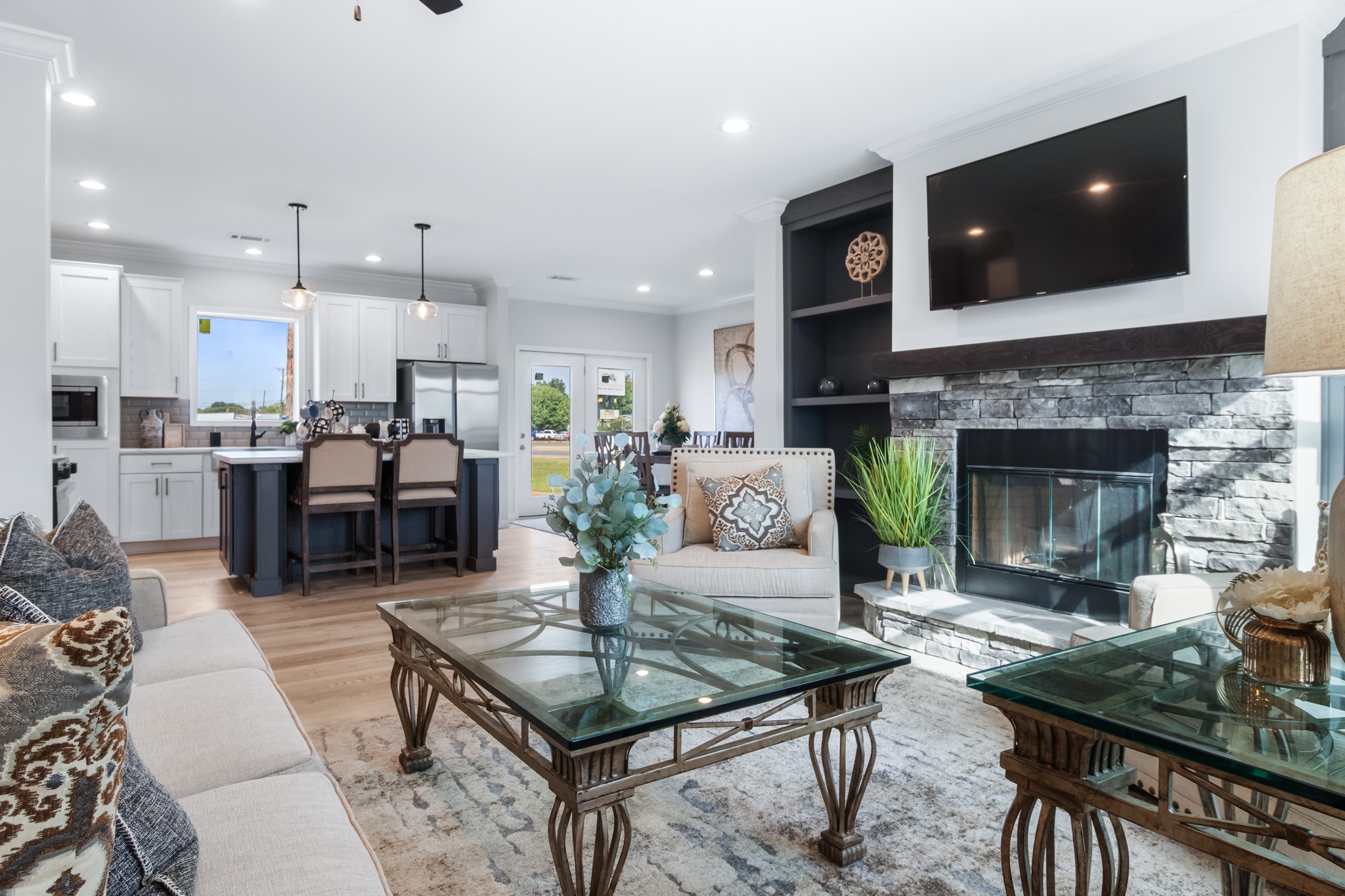 Chic living room with stone fireplace, mounted TV, and glass tables. Open kitchen has pendant lights, white cabinets, and a breakfast bar. Cozy, elegant vibe.