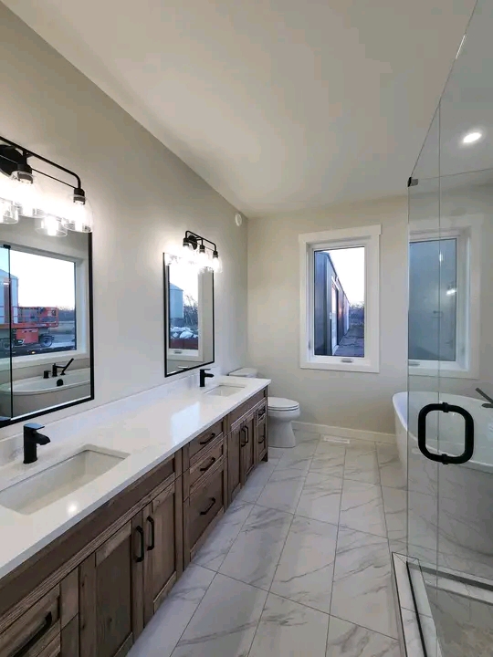 Modern bathroom with a double vanity, wooden cabinets, and black fixtures. Large mirrors reflect the natural light from a window. Marble tiles add elegance.