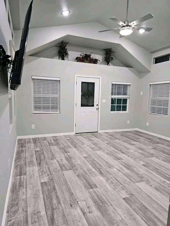 A minimalist room with gray wooden floors, a white door with frosted glass, and two windows with blinds. Plants sit on a ledge above, and a ceiling fan adds brightness.